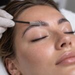 Gloved hand brushing a client's brows upward with a spoolie during a brow lamination