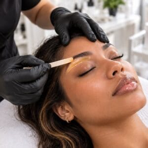 Esthetician in black gloves applying warm wax under a client's brow with a wooden spatula